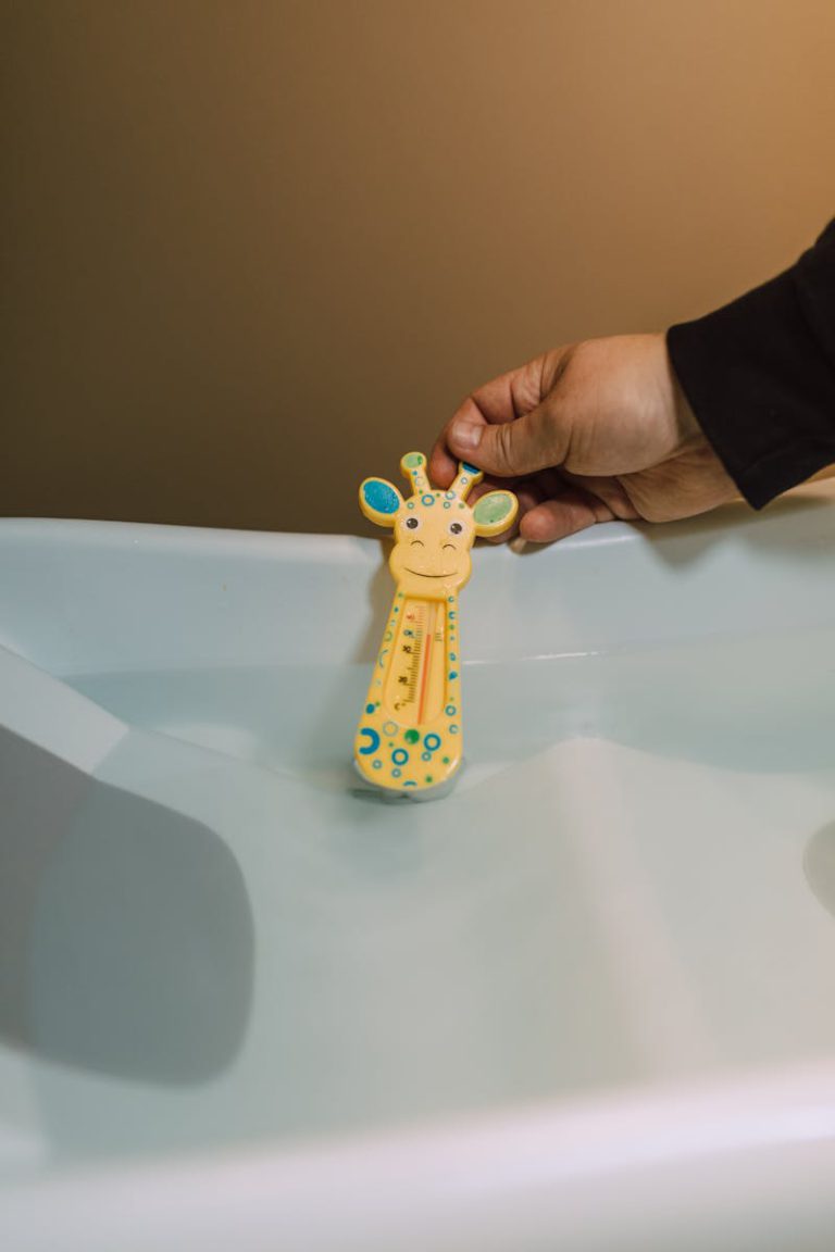 A hand holds a giraffe-shaped bath toy thermometer in a water basin.