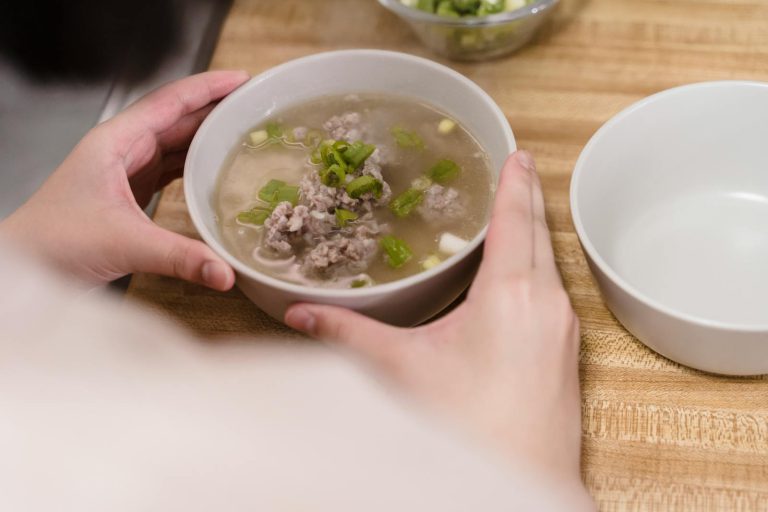 Close-up of a bowl of pork soup with green onions held indoors, highlighting warmth and comfort.