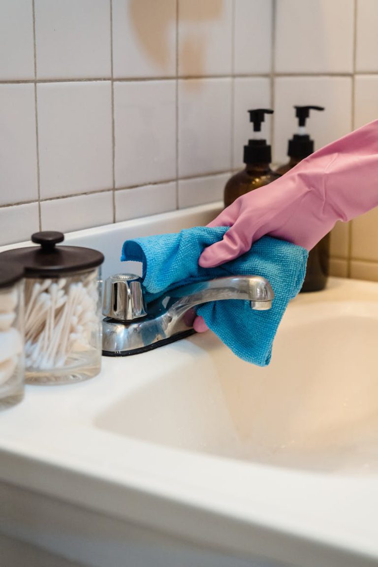 A gloved hand cleans a bathroom sink faucet with a blue cloth for hygiene.